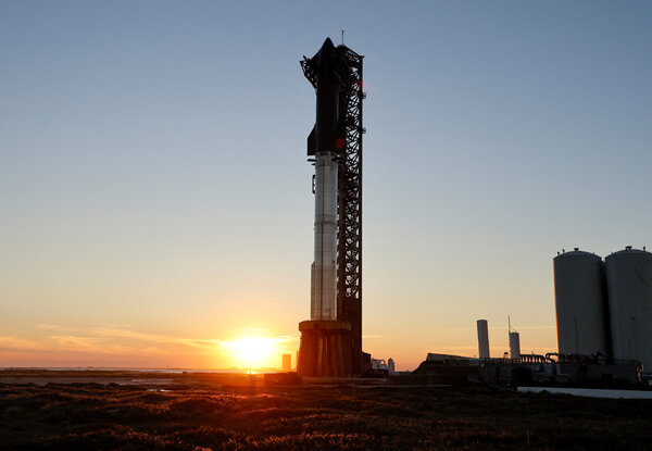 The setting sun sits just above the horizon as the enormous Starship rocket waits at the launchpad against a clear sky.